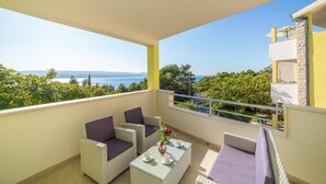 Ein Balkon mit Korbmöbeln, einem Tisch mit Tassen und einer Vase, und Blick auf das Meer und Grünpflanzen.