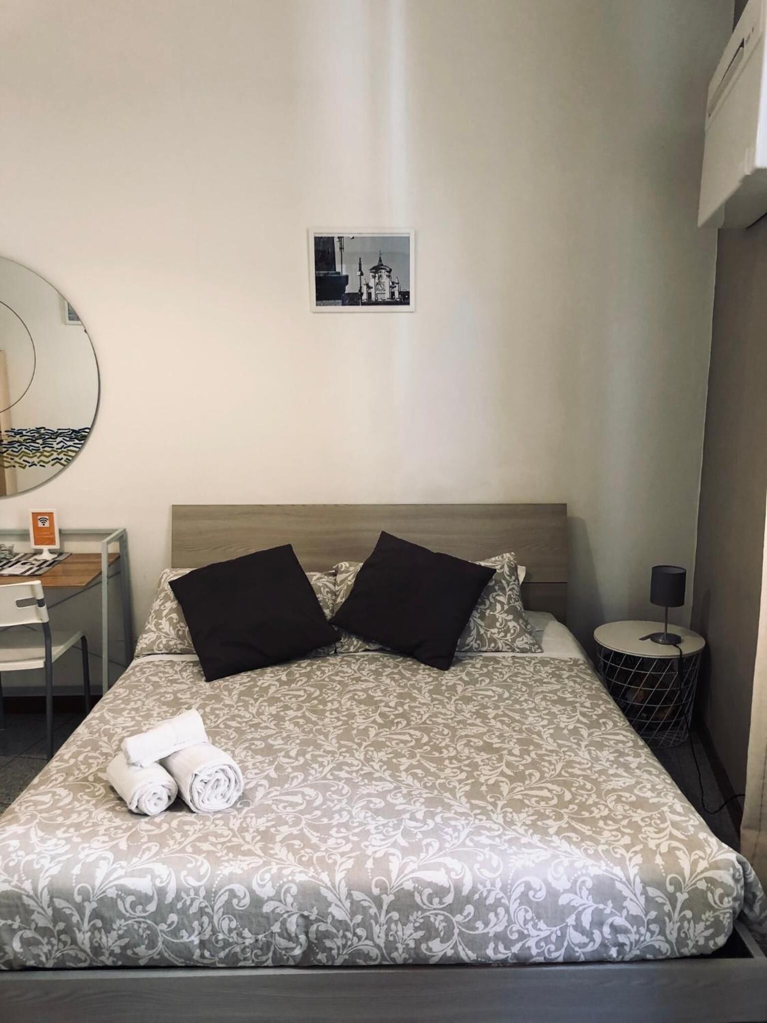 Una cama bien hecha con ropa de cama estampada, una mesita de noche con lámpara, un escritorio con computadora y una silla, y un espejo montado en la pared.