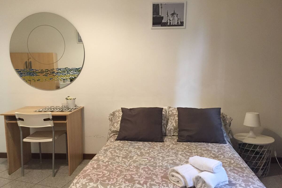A neatly arranged bedroom with a bed, a desk, a chair, a round mirror, a small table with a lamp, and a framed picture on the wall.
