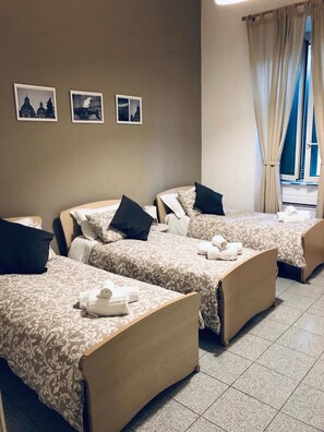 A hotel room with three beds, each with a patterned bedspread and pillows. There are three framed pictures on the wall, a window with curtains, and a tiled floor. - Domus Santa Caterina (Rome)