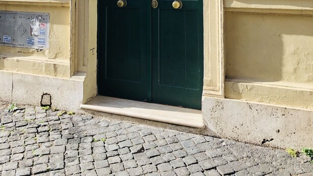 A double door with a metal grille and a small window above it, set in a beige wall with a number plate '49' visible. - Domus Santa Caterina (Rome)