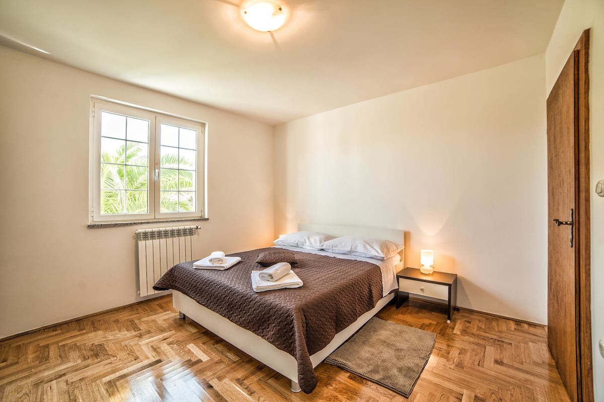 A bedroom with a bed, a nightstand, a lamp, a window, and a radiator.