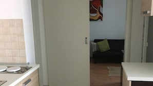 Ett kök med en vit bänkskiva, en diskho och ett skåp med lådor. Det finns en dörr som leder till ett vardagsrum med en soffa och en tavla på väggen.