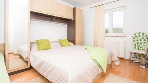 Ett sovrum med en säng, en garderob i trä, ett element, ett fönster med gardiner och en stol med en kudde med prickmönster.