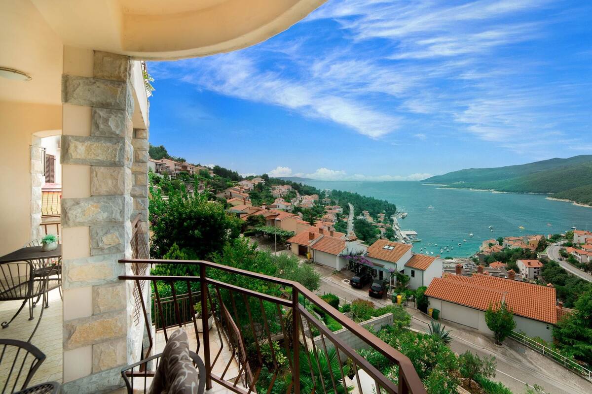 Varanda com parede de pedra, com vista para uma vila costeira com casas e barcos.