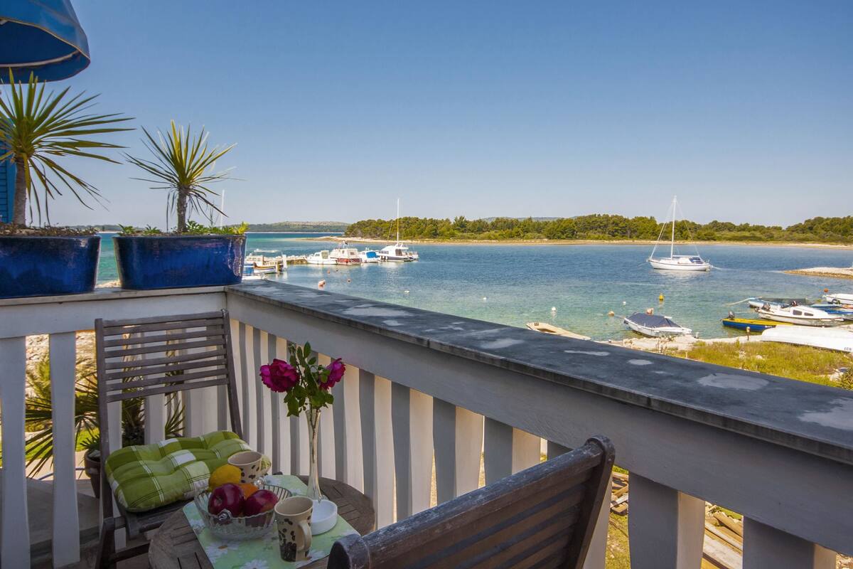 A balcony with a table set for two, overlooking a marina with boats and a clear view of the sea.