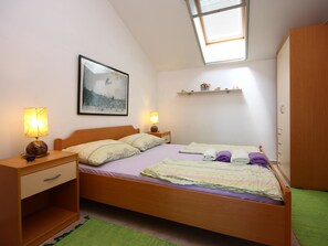 Ein Schlafzimmer mit einem Bett, zwei Nachttischen, einem kleinen Tisch, einem grünen Teppich, einem Fenster mit Jalousien, einem Wandregal mit Dekoration und einem gerahmten Bild an der Wand.