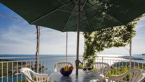 A round table with a patterned tablecloth, surrounded by white chairs, under a green umbrella, overlooking a scenic coastal view. - Apartments Nikica Primosten (Primosten)