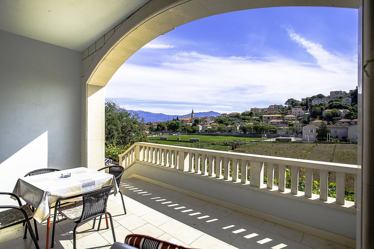 A balcony with a table and chairs, overlooking a town with buildings and mountains in the distance.