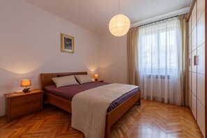 Een slaapkamer met een houten bed, twee nachtkastjes met lampen, een hanglamp, een ingelijst schilderij aan de muur, en een raam met dunne gordijnen.
