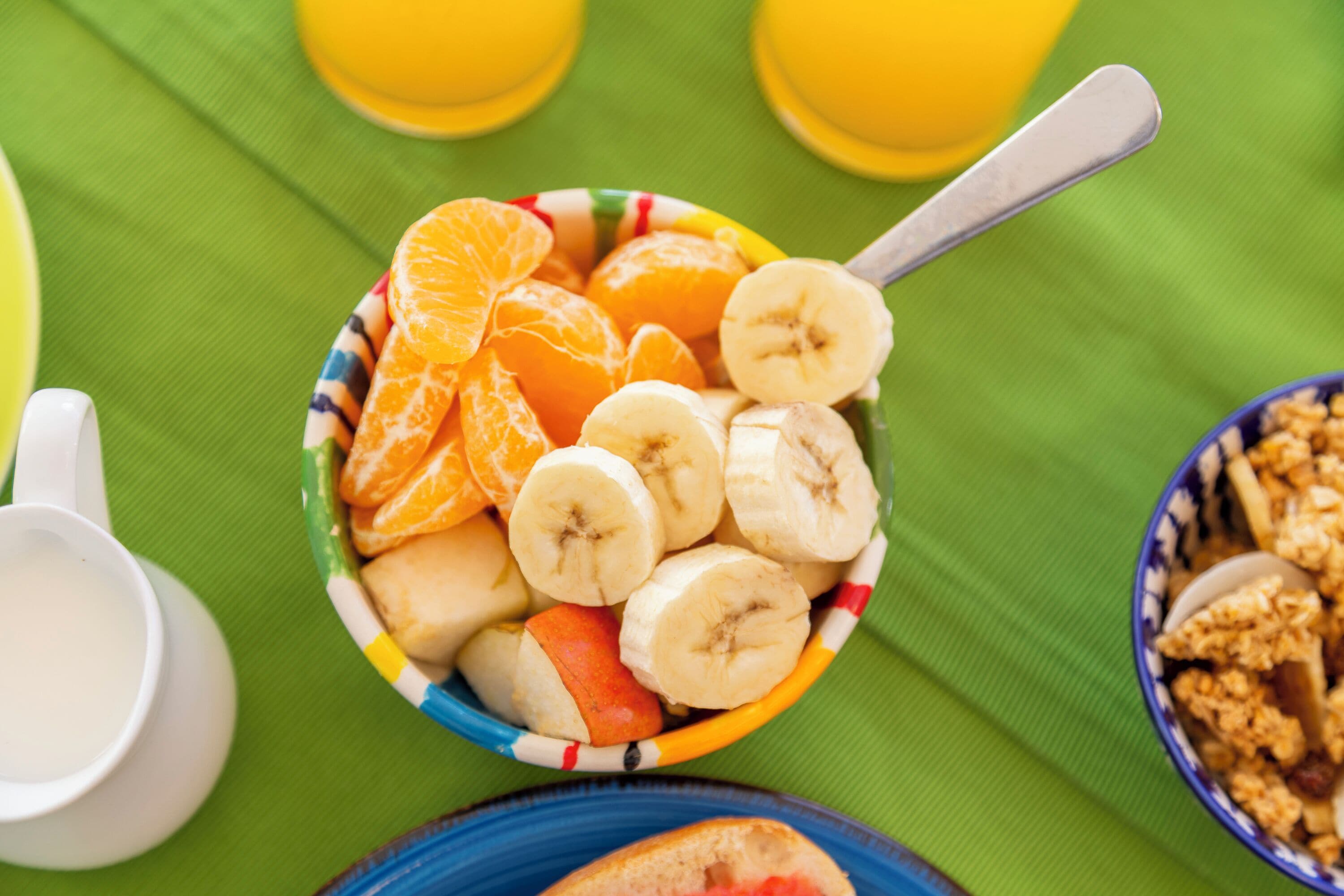 Un tazón de fruta con plátanos y naranjas, una cuchara y un tazón de granola sobre un mantel verde.