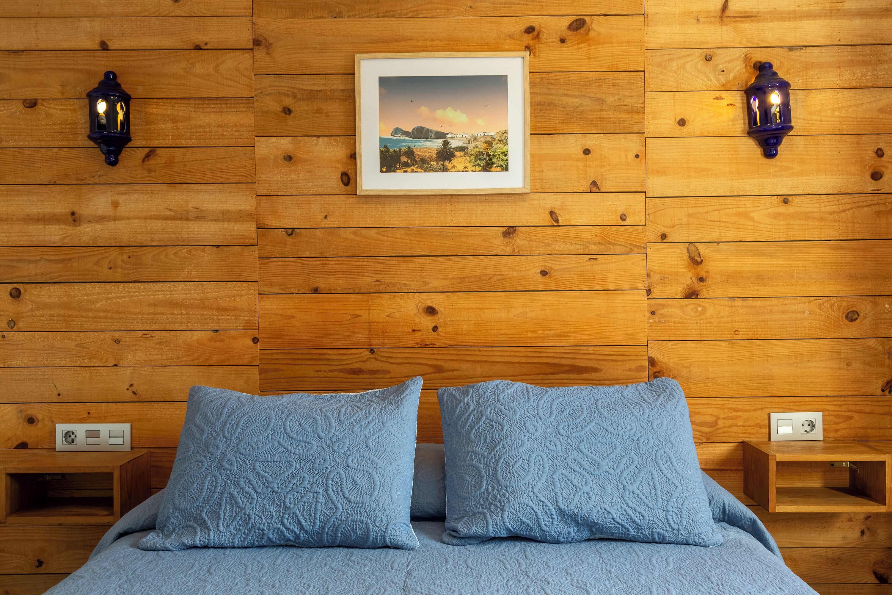 Una habitación con paneles de madera, una cama, dos almohadas a juego, un cuadro enmarcado y dos faroles fijados a la pared.