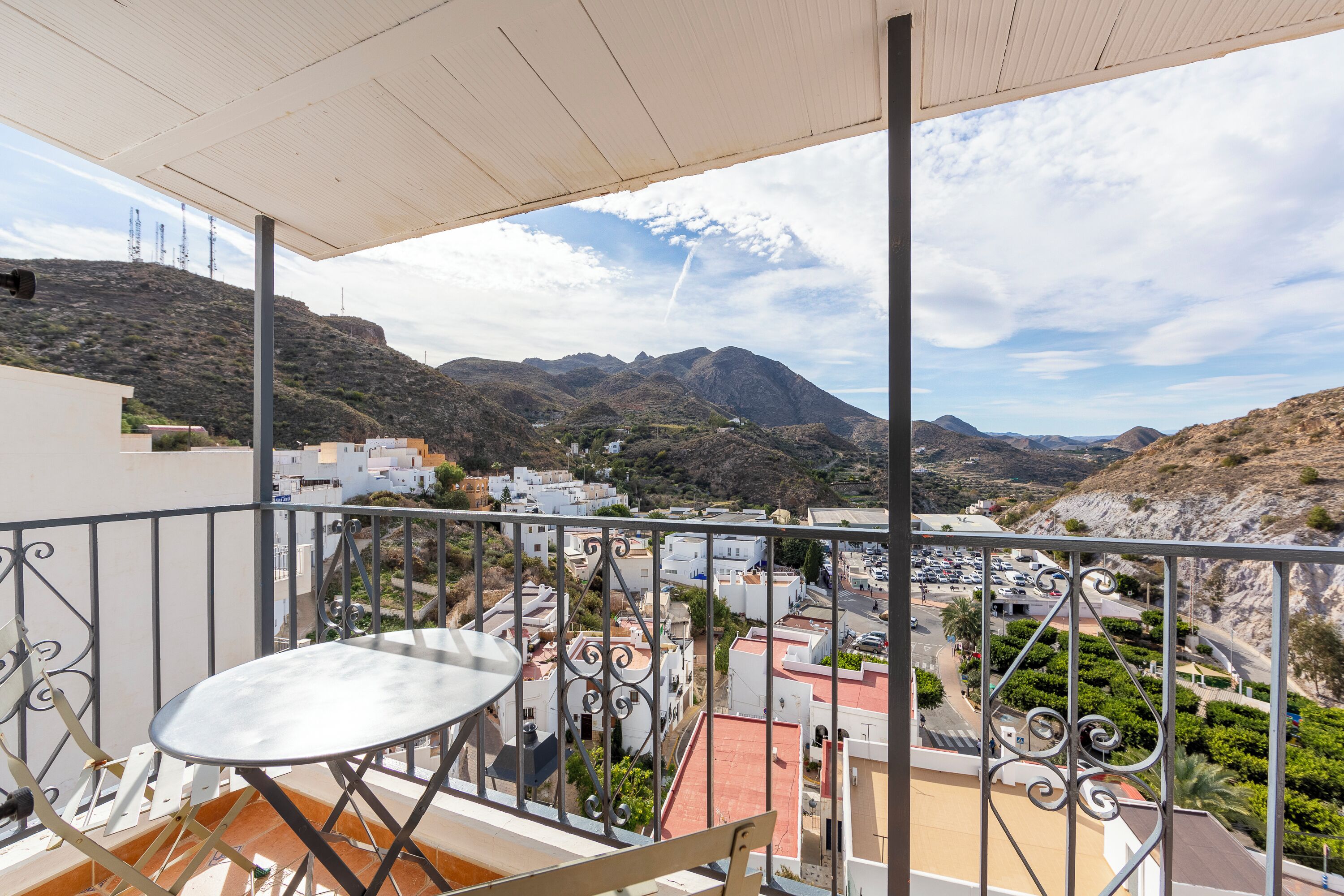 Un balcon doté d’une balustrade en métal, une table blanche et une vue sur une ville, avec des bâtiments et des montagnes en arrière-plan.