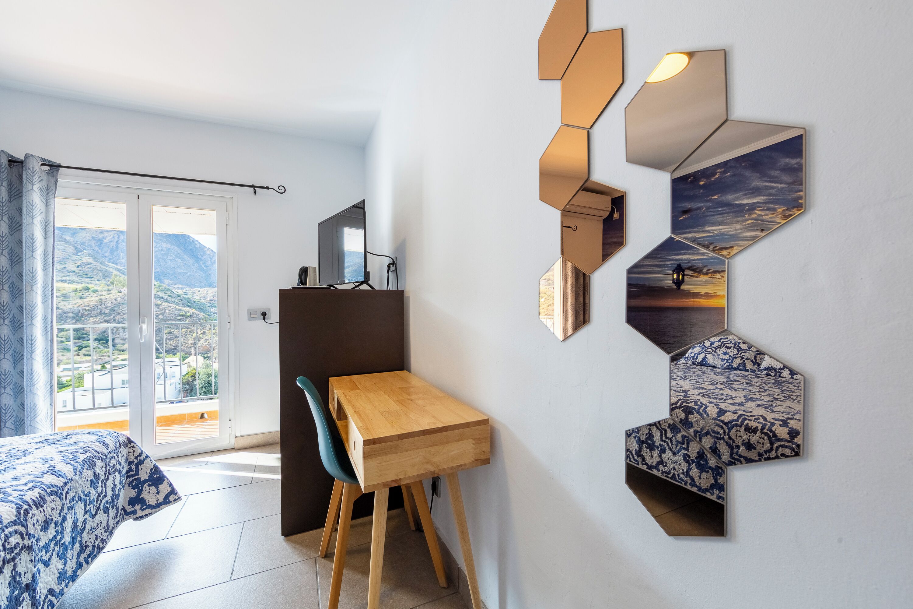A modern room with a wooden desk, a chair, and a bed with patterned bedding. There is a balcony with a view of the mountains.