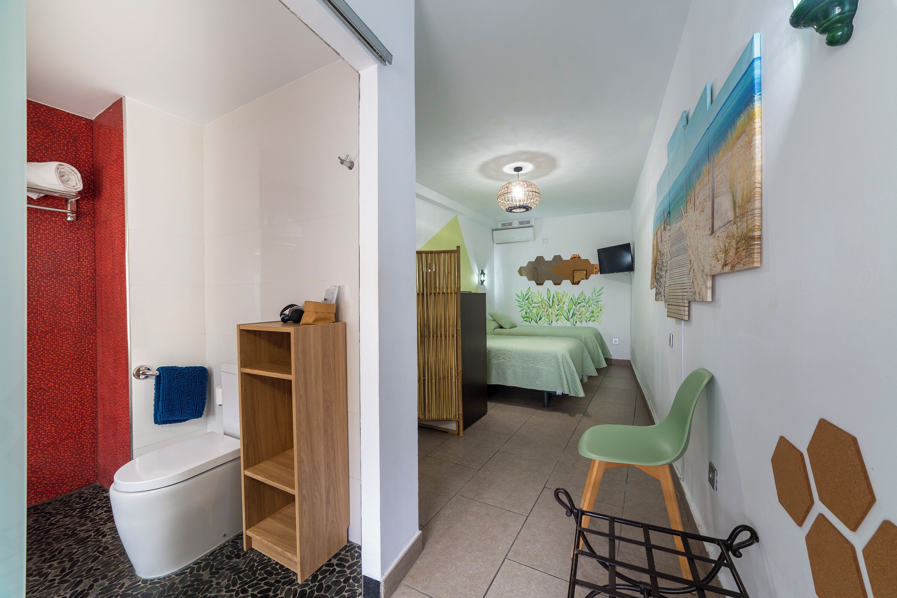 A hotel room with a toilet, a wooden shelf, a green chair, a bed with a green bedspread, and a wall with a decorative painting.