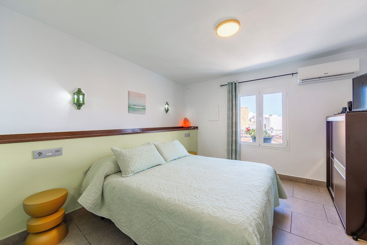 Un dormitorio con cama, mesita de noche, lámpara de pared y ventana con vistas al exterior.