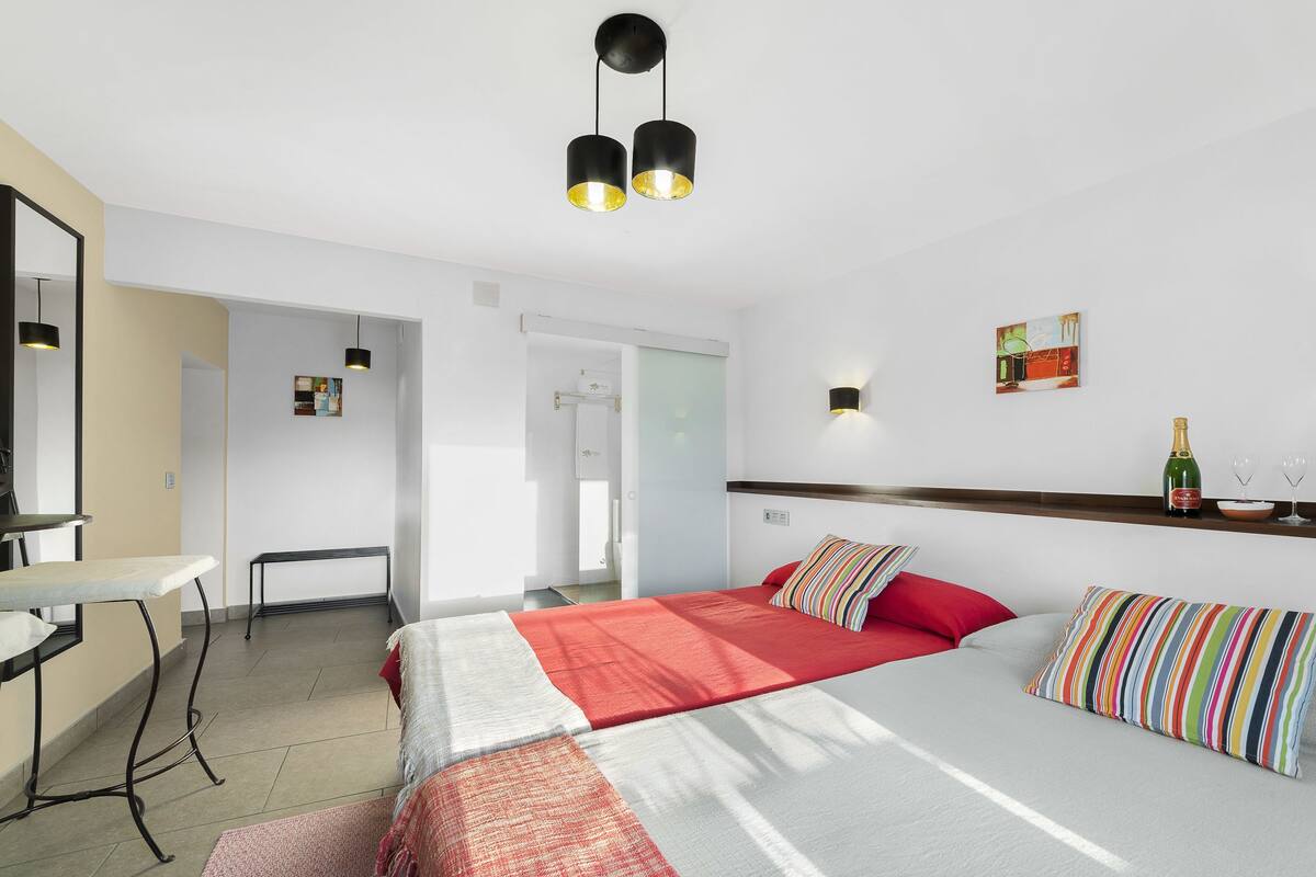 Habitación de hotel con cama, escritorio, silla, espejo, lámpara fijada a la pared y una botella de champán en un estante.