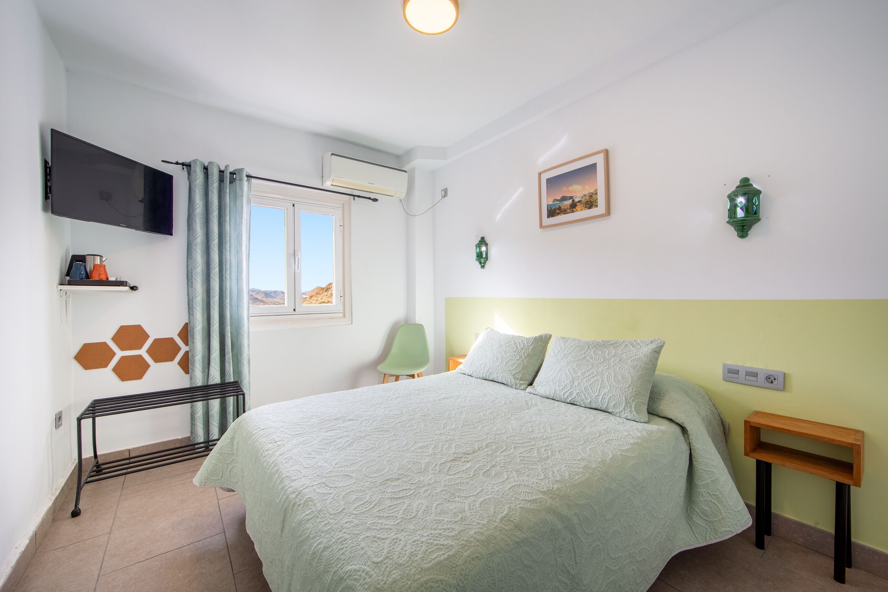 Un dormitorio con cama, televisión, ventana, silla y una mesita.