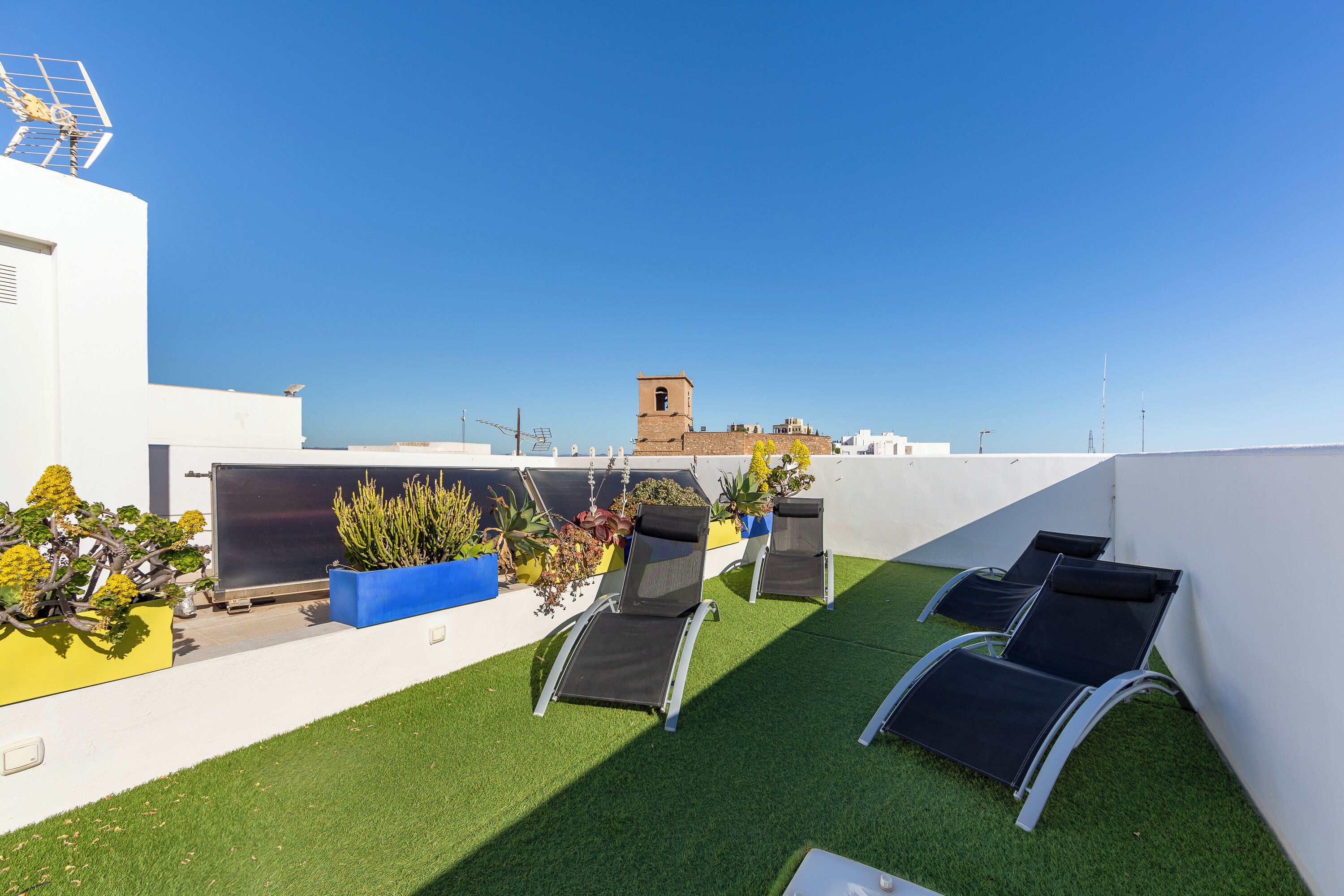 Terraza en la azotea con césped artificial, dos sillones de descanso, plantas en macetas y una vista despejada de una torre distante.