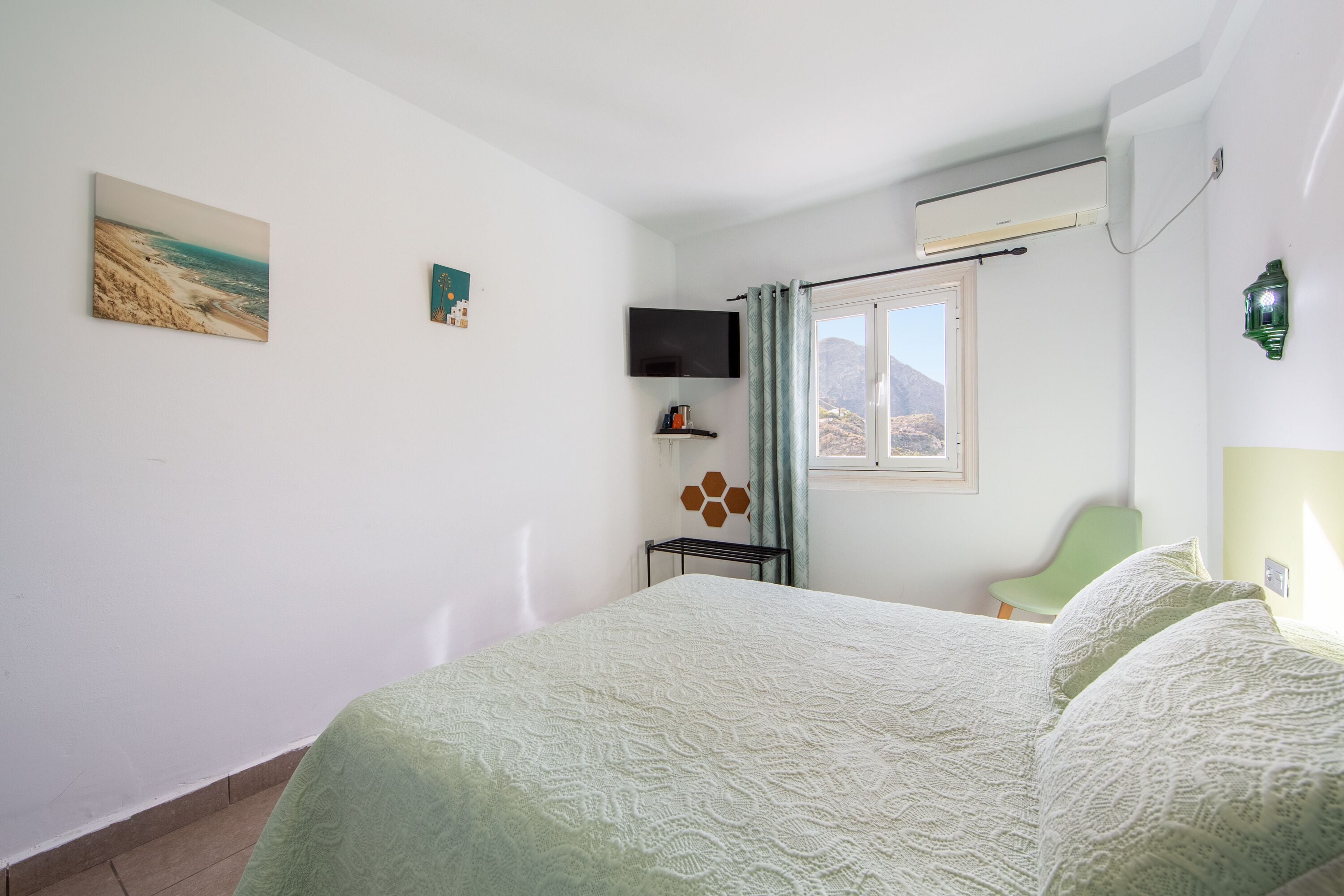 Ein Schlafzimmer mit einem Bett, einem an der Wand befestigten Fernseher, einem Fenster mit Vorhängen und einer Wand-Klimaanlage.
