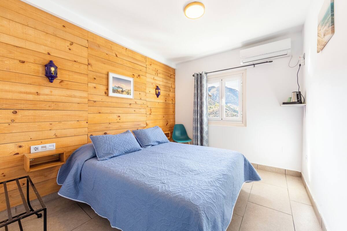 Un dormitorio con pared de madera, una cama con ropa de cama azul, una silla verde y una ventana con vistas a montañas.