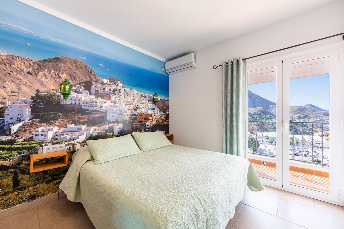 Un dormitorio con una cama grande, un mural de un pueblo costero y un balcón con vistas al pueblo y a las montañas.