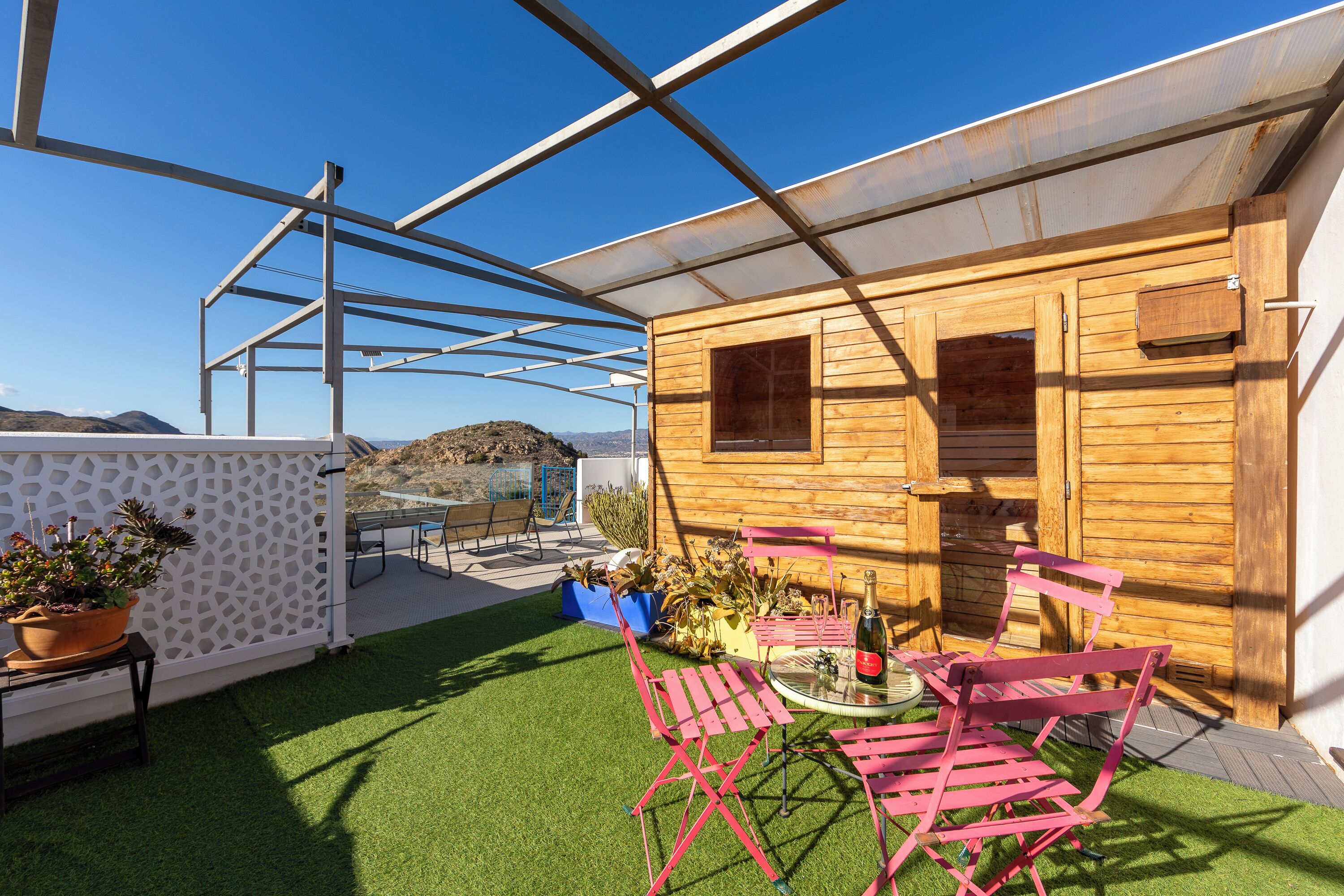 Una cabaña de madera con patio y techo de vidrio, césped artificial verde y sillas de metal rosa.
