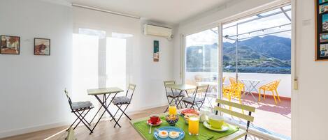 Un comedor luminoso con una mesa lista para el desayuno, un balcón con sillas y vistas a la montaña.