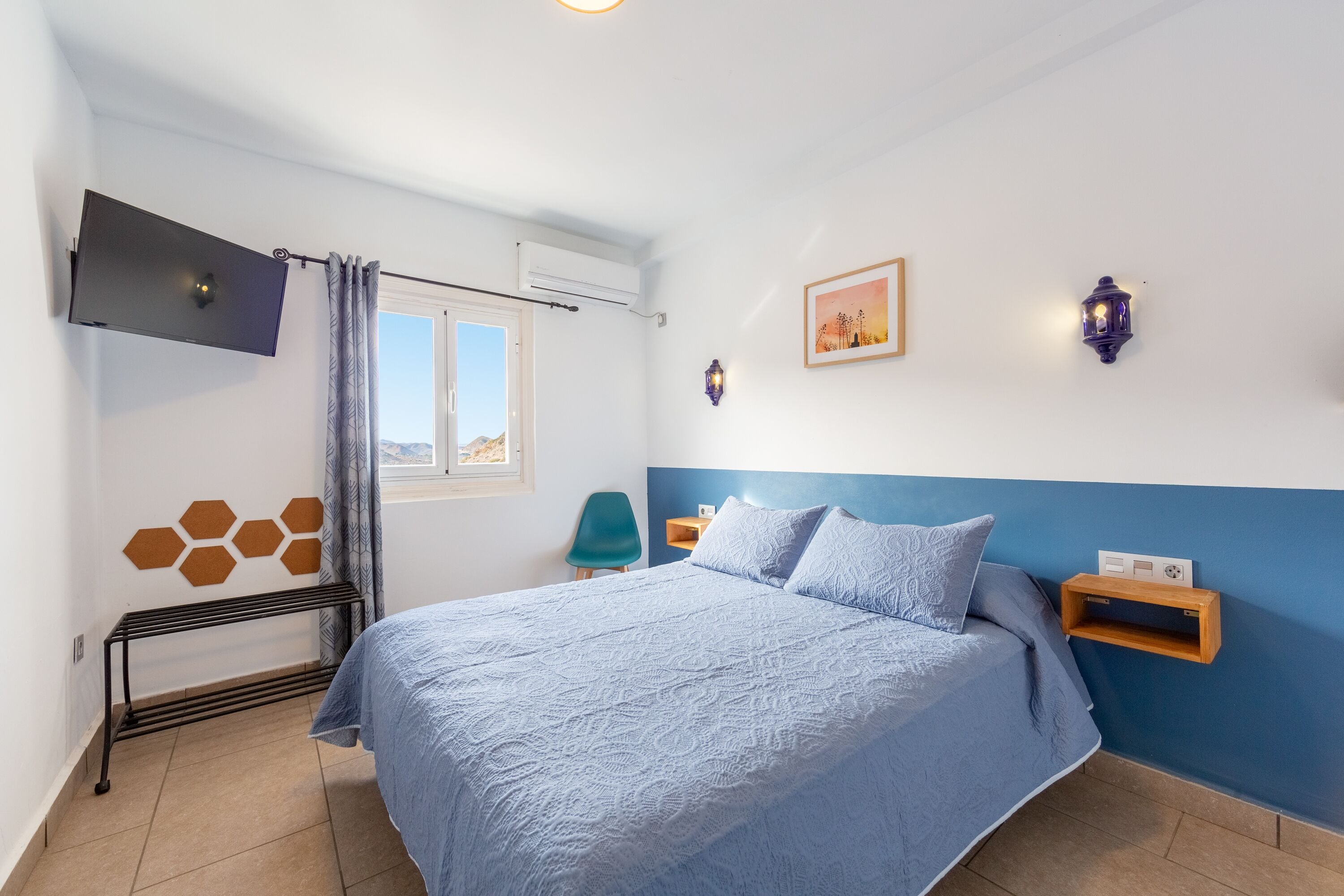 Een slaapkamer met een bed, een aan de muur bevestigde televisie, een raam met gordijnen, een wandplank en een fotolijst.