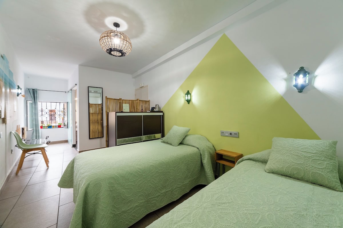 Una habitación con dos camas, un escritorio, una silla, un ventanal con cortinas y un rincón americano.