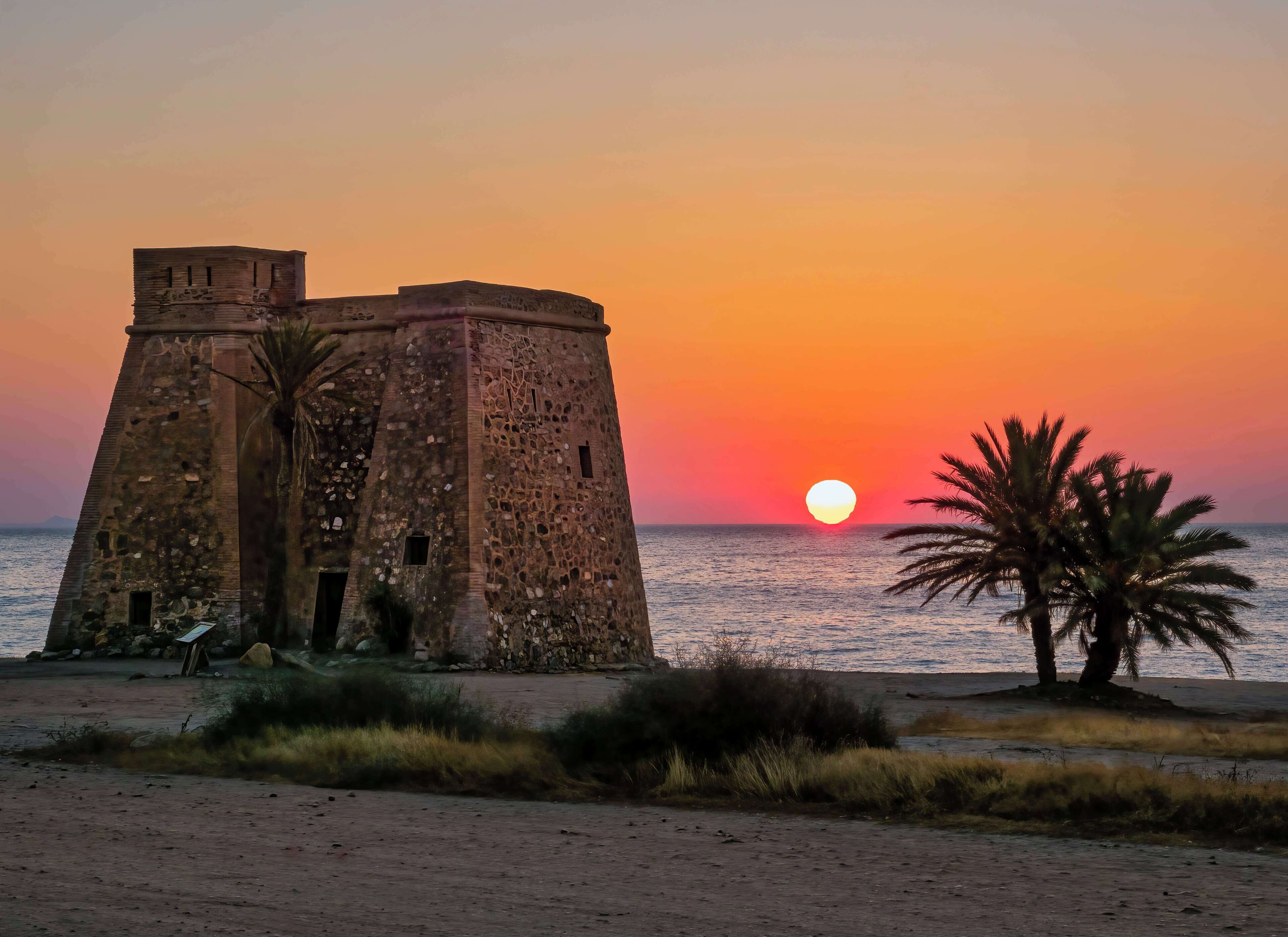 A coastal sunset with a historic stone tower and palm trees.