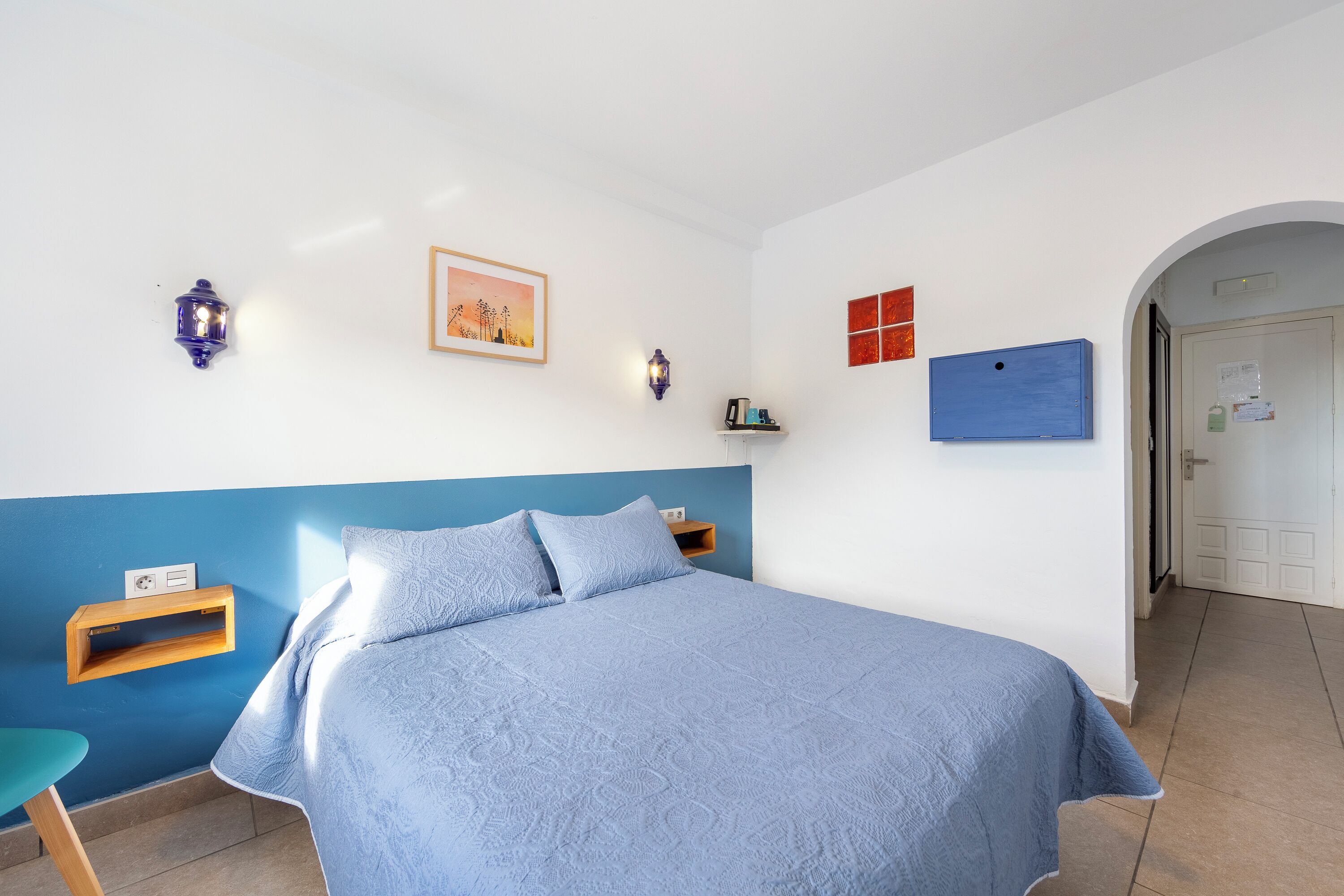 A bedroom with a bed, a blue wall, a wooden shelf, a picture, a wall-mounted light, and a door.
