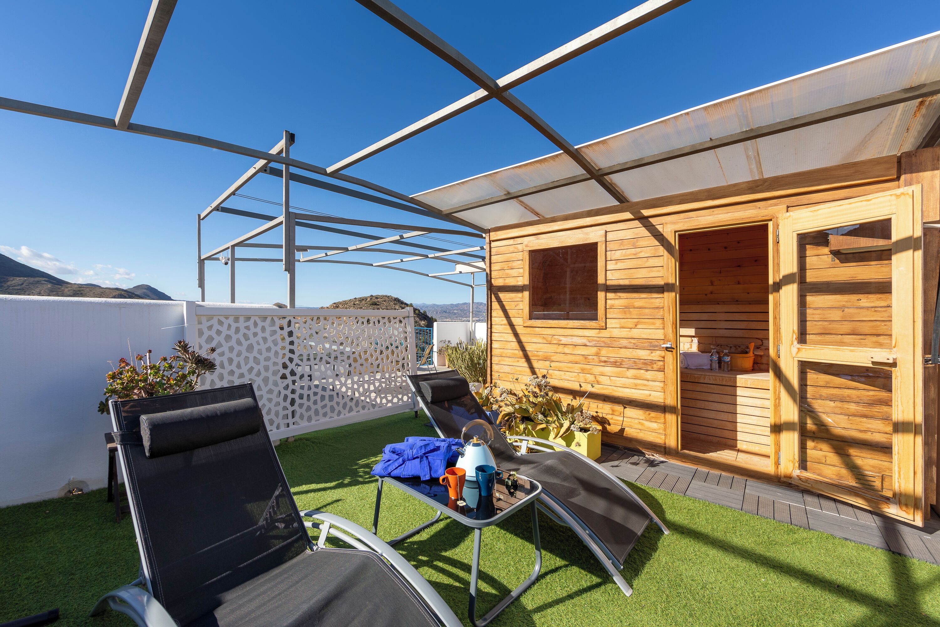 A covered outdoor area with two lounge chairs, a table with a kettle and mug, and a wooden building with a door.