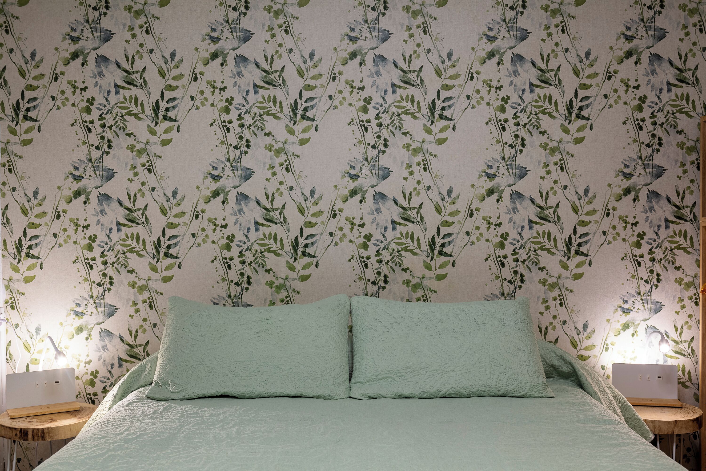 A bedroom with a floral wallpaper, a bed with two pillows, and a laptop on a bedside table.