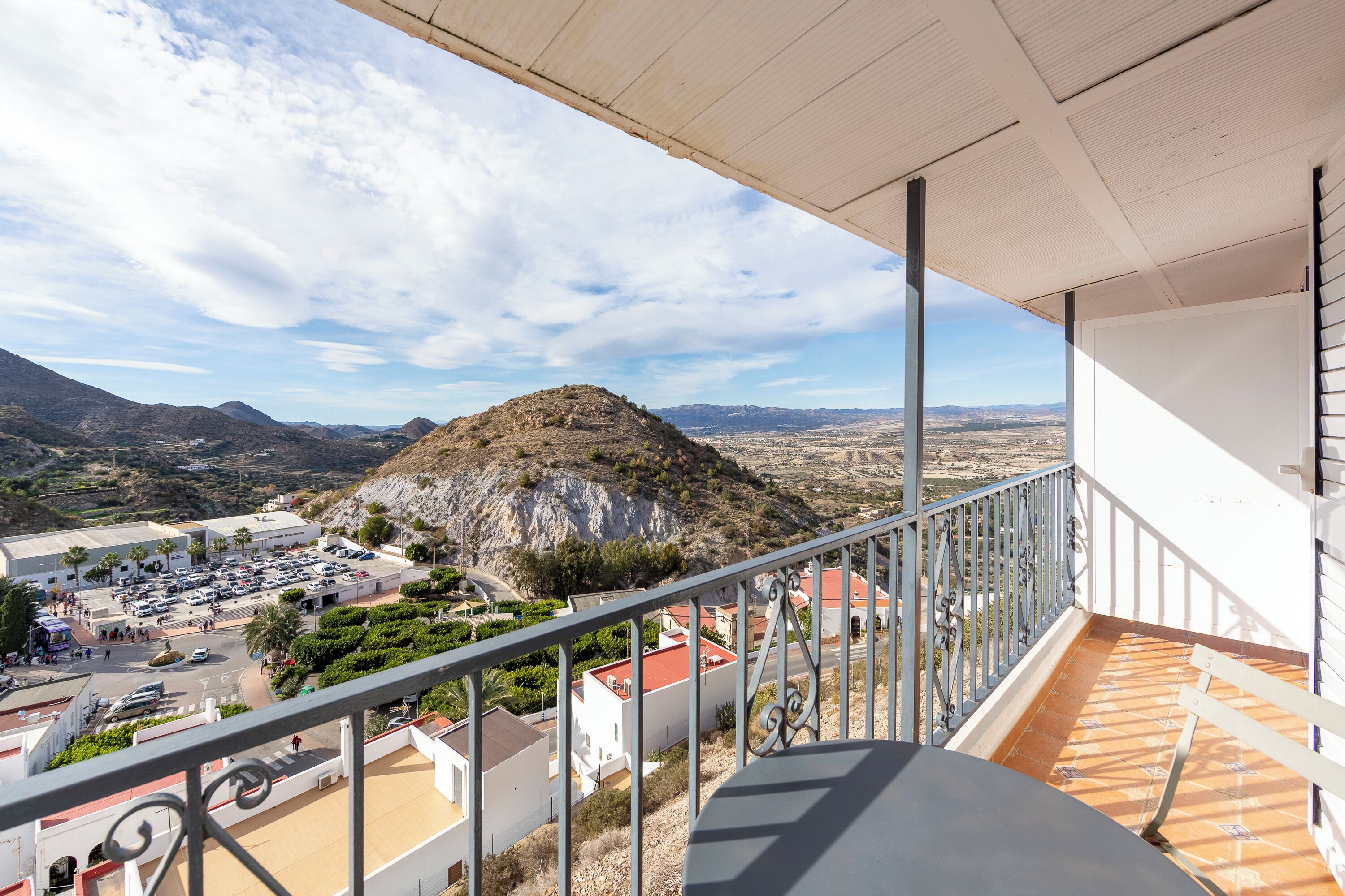 A balcony with a view of a mountainous landscape, a parking lot, and buildings with red roofs.