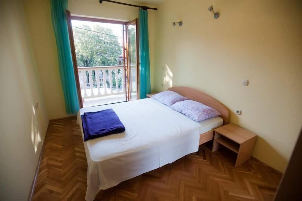 Ein Schlafzimmer mit einem Bett, einem kleinen Nachttisch, einem Fenster mit Vorhängen und einem Balkon.
