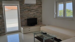 A modern living room with a glass coffee table, a flat-screen TV on a stone wall, a sliding glass door leading outside, and a window with a view of trees. - Apartments MIA Bilo 887 (Primosten)