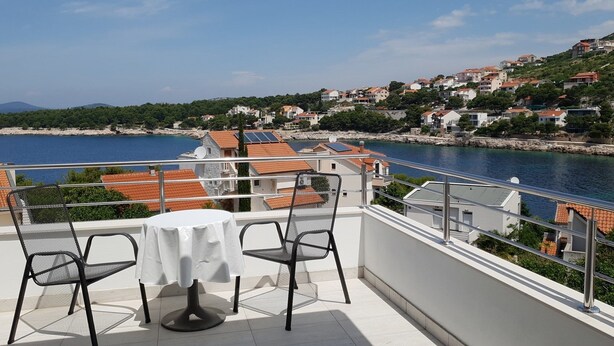 A balcony with a table and chairs overlooking a coastal town with red-roofed buildings and a body of water. - Apartments MIA Bilo 887 (Primosten)