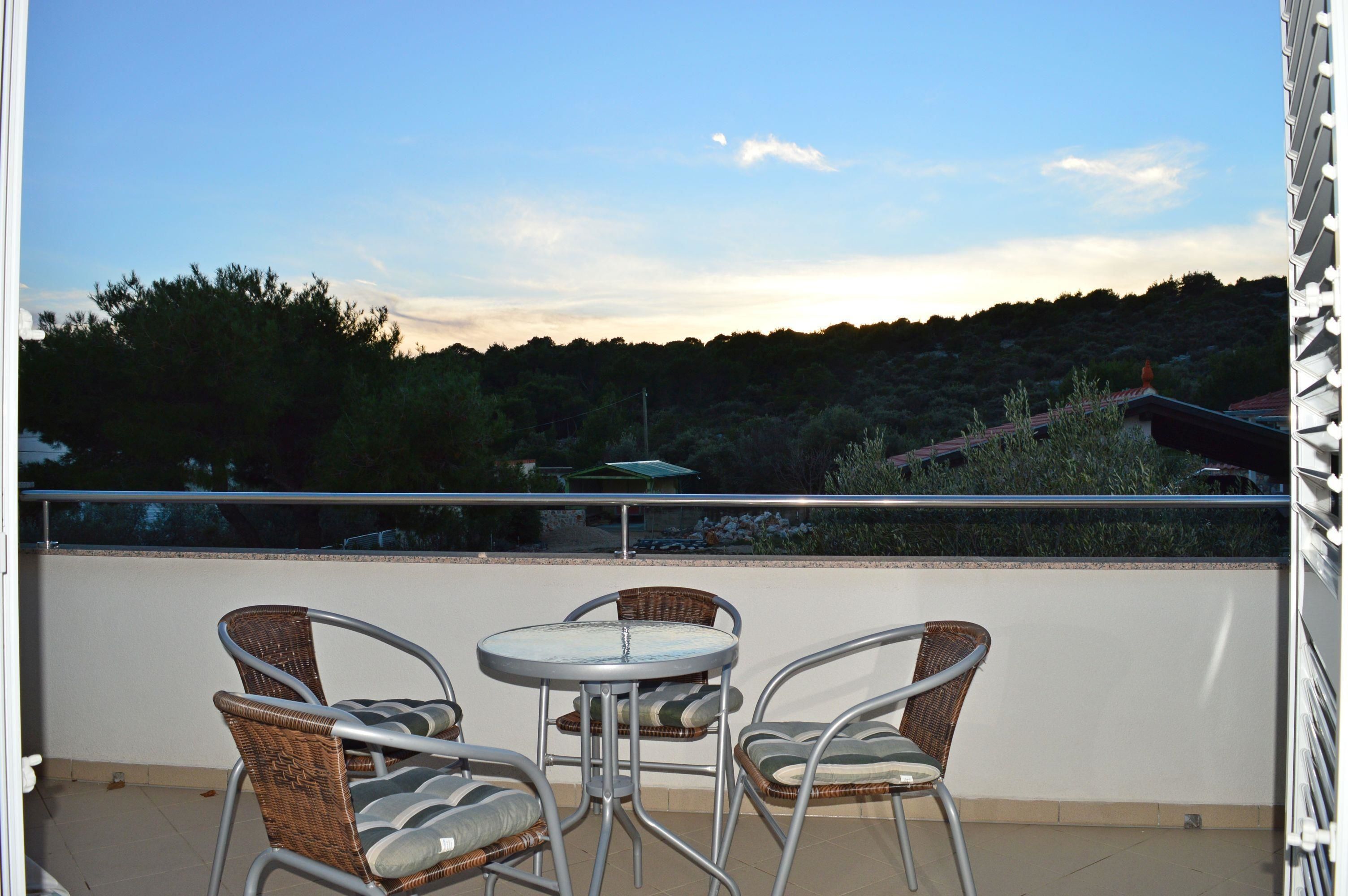 A balcony with wicker chairs and a small round table, overlooking a hilly landscape with trees and a distant building.