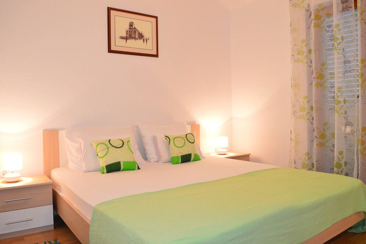 A neatly made bed with a green blanket and two pillows, a bedside table with a lamp, and a framed picture on the wall.