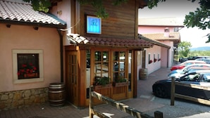 A rustic building with a tiled roof, wooden doors, and a small porch area. There are cars parked outside and a stone wall to the left. - Fattoria Carsica Bajta (Sgonico)
