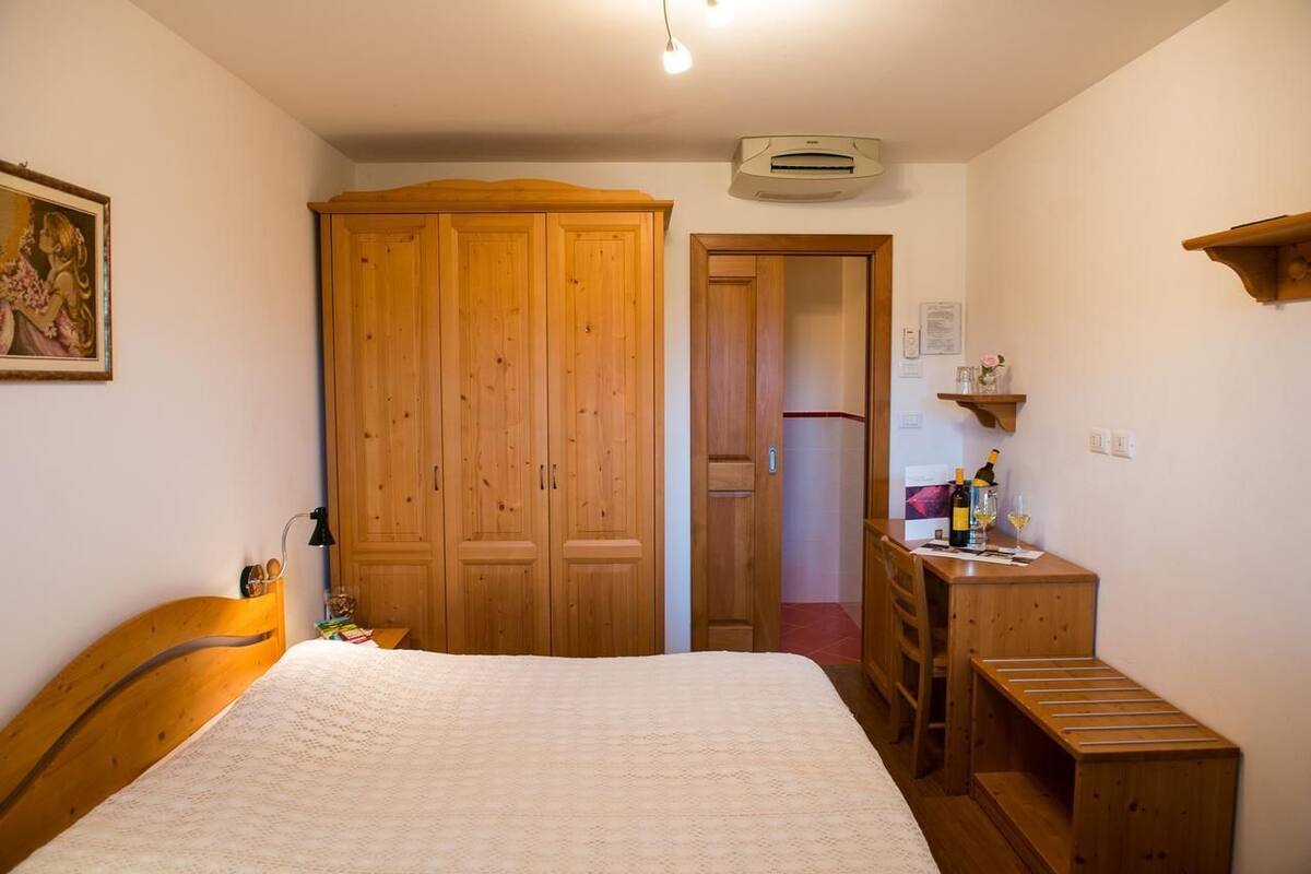 A bedroom with a wooden wardrobe, a bed with a white blanket, a desk with a chair, and a small shelf with items.