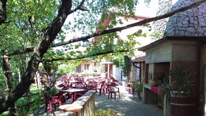 An outdoor patio with red plastic chairs and tables, surrounded by trees and a stone building. - Fattoria Carsica Bajta (Sgonico)