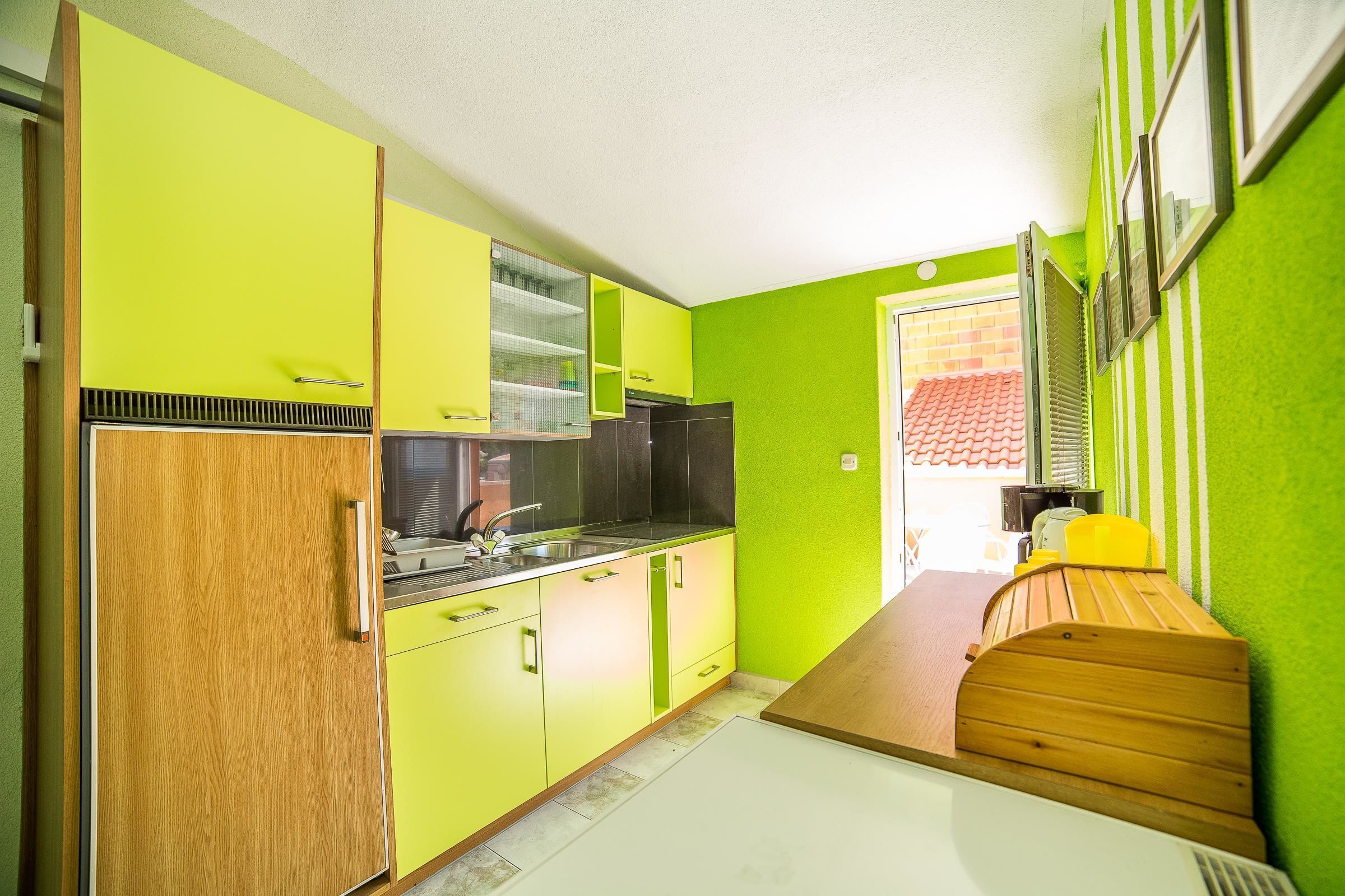 A kitchen with green cabinets, a wooden bench, and a window with a red-tiled roof visible through the door.