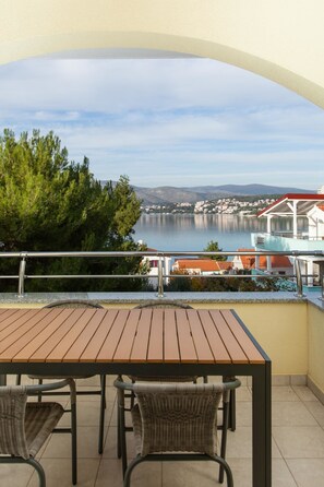 A balcony with a wooden table and wicker chairs, overlooking a lake with buildings in the background.