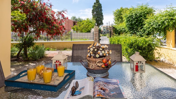 A covered outdoor seating area with a table set for breakfast, including orange juice, pastries, and a bowl of fruit. - Maria's Countryside House (Corfu)