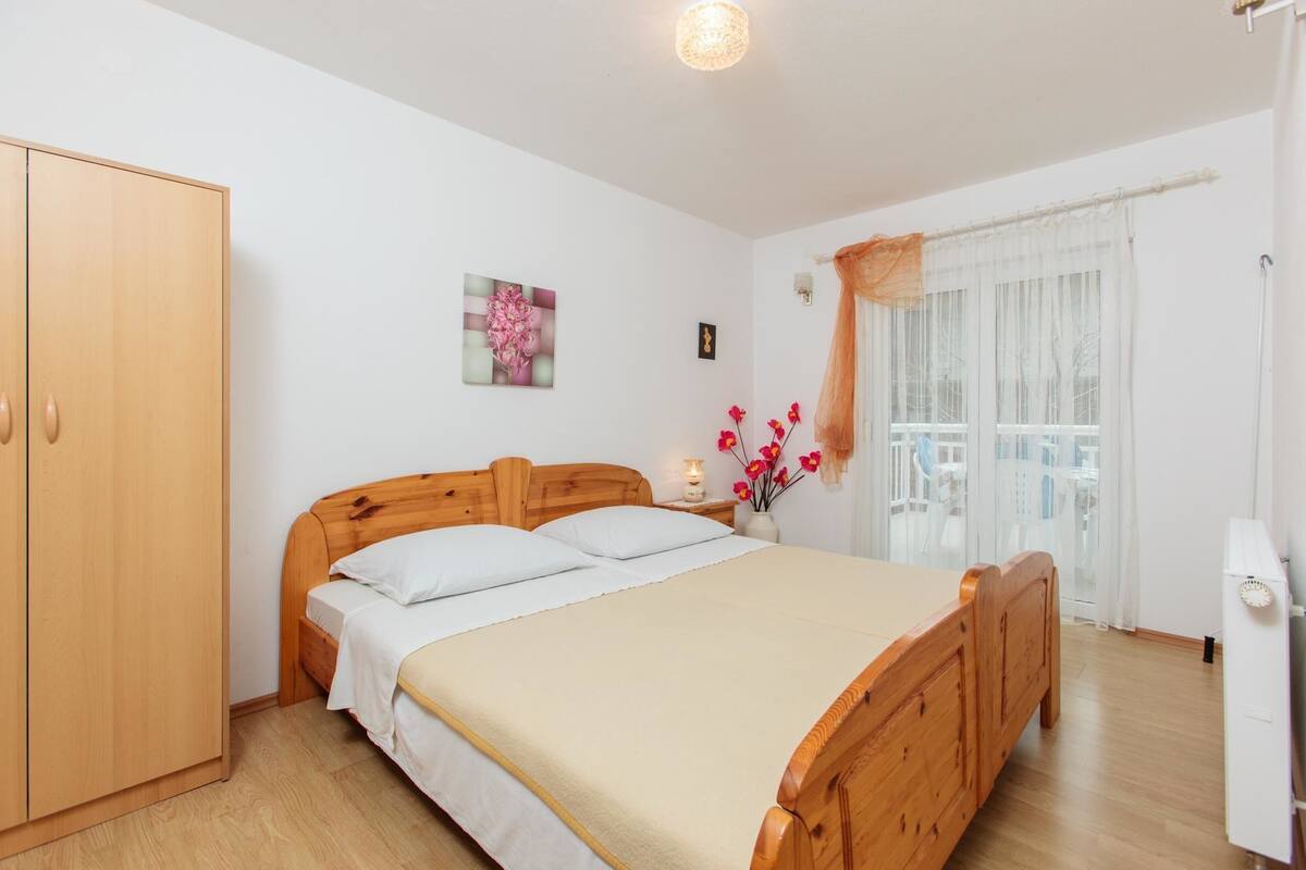 A bedroom with a wooden bed, a wardrobe, a nightstand with a lamp, a window with sheer curtains, and a wall-mounted picture.