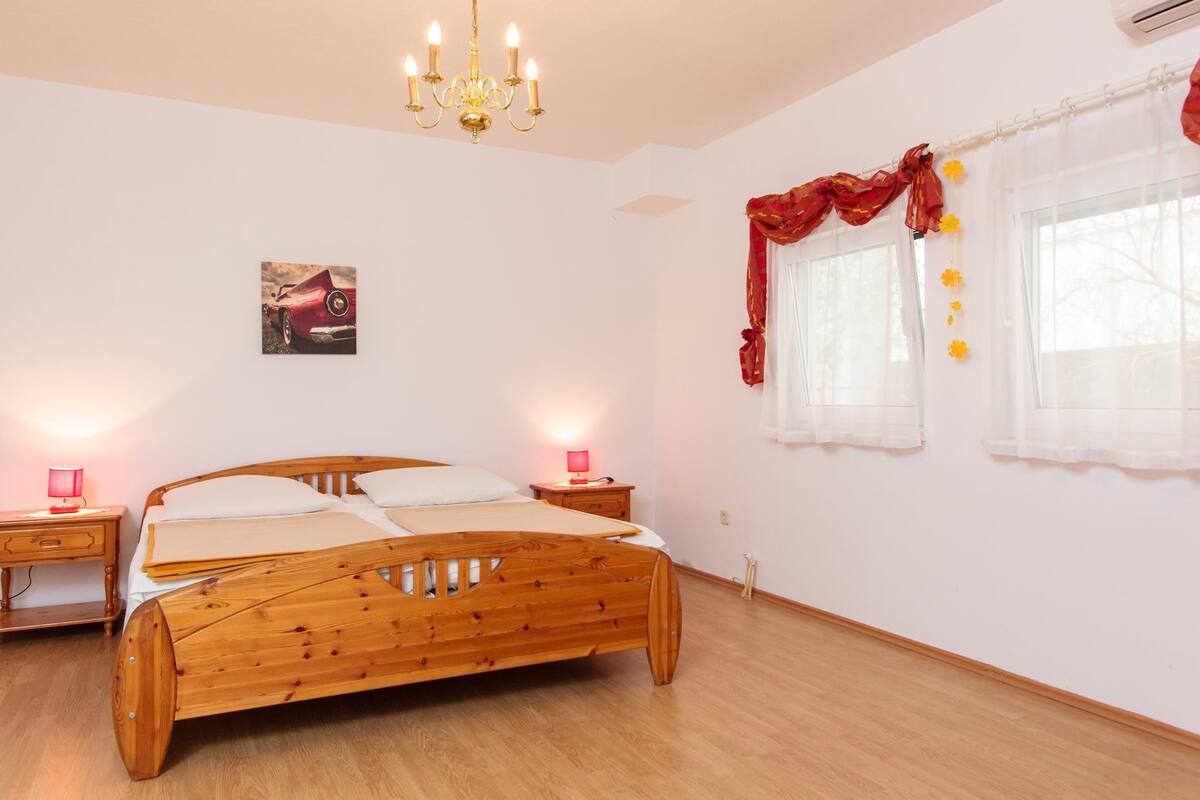 A bedroom with a wooden bed, two bedside tables with lamps, a chandelier, a painting on the wall, and a window with curtains.