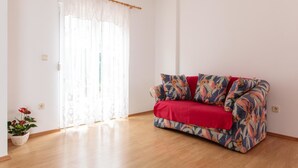 A room with a wooden floor, a floral-patterned sofa with a red cover, a potted plant, and sheer curtains on a window. - Apartments Villa Dane (Podstrana)