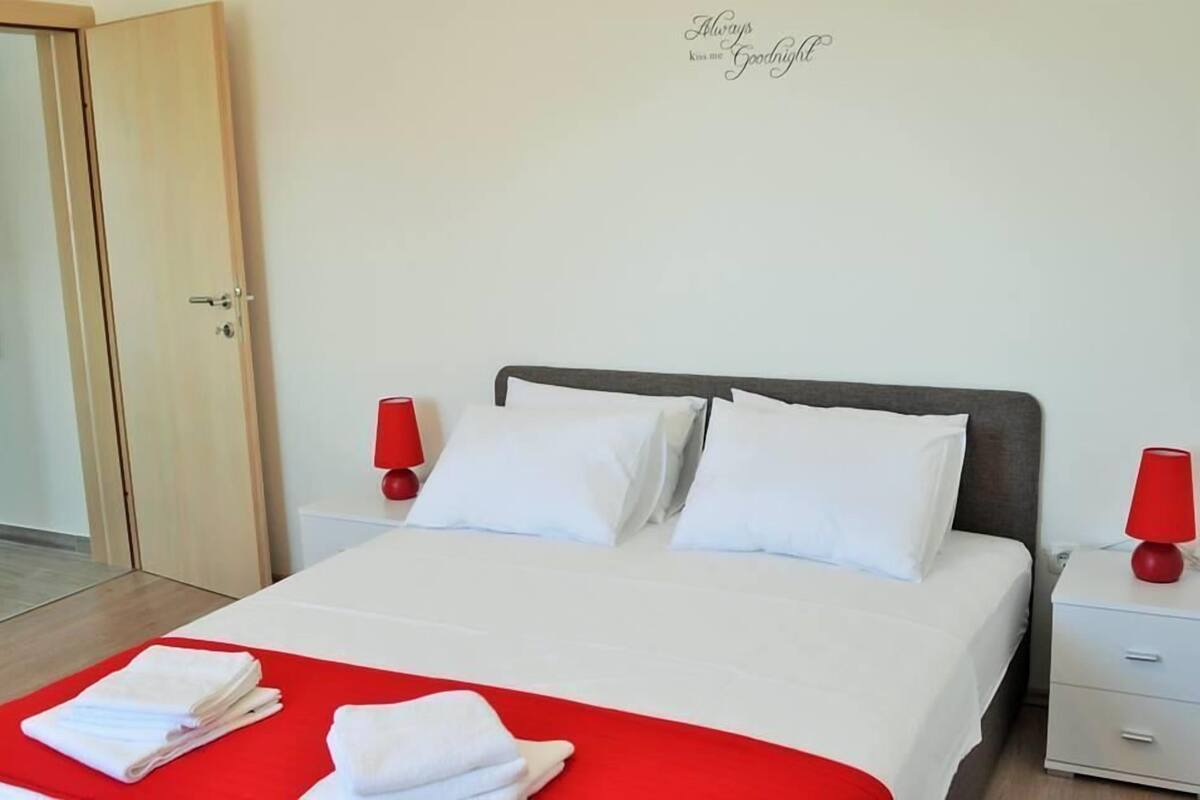 A neatly made bed with white linens and a red accent, two bedside tables with red lamps, and a wall sign.